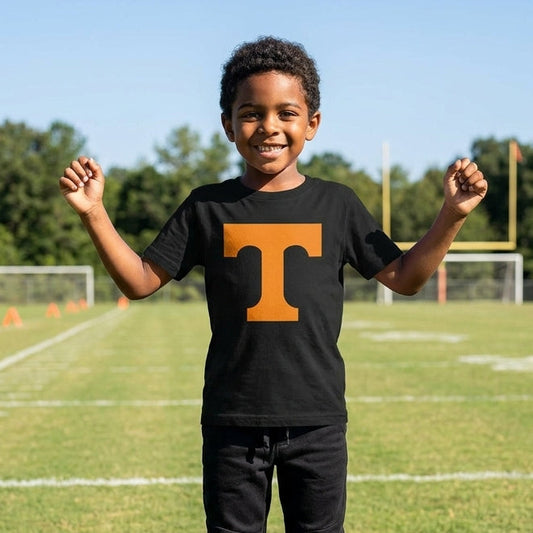 A young boy stands on a grassy football field wearing a black short-sleeve t-shirt with a large orange block letter 'T' on the chest, paired with black sweatpants. The background shows a sunny outdoor sports field with goalposts, trees, and a blue sky.
