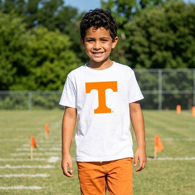 A young boy stands on a grassy football field wearing a white short-sleeve t-shirt with a large orange block letter 'T' on the chest, paired with orange pants.