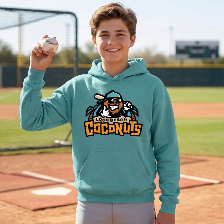 A smiling young man stands on a baseball field wearing a teal hoodie with a custom logo graphic on the front, holding a baseball up in one hand.