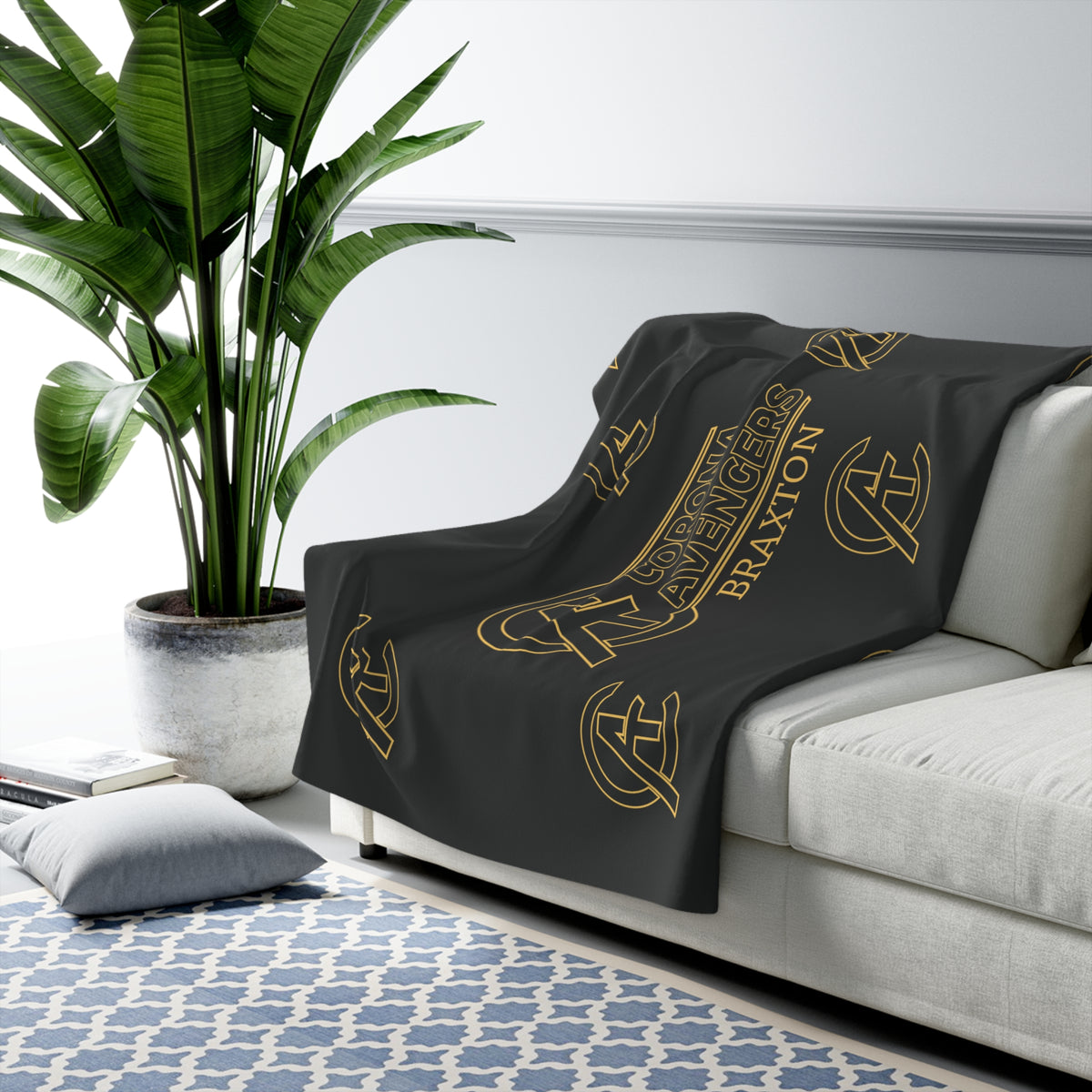 A dark charcoal throw blanket with gold printed logos and the word 'BRAXTON' draped over a light gray sofa in a modern living room setting with a potted plant and patterned rug.