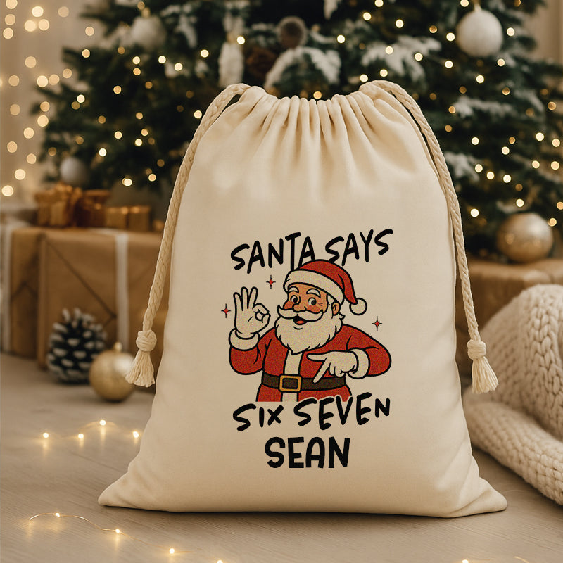 Beige drawstring bag with Santa Claus graphic and text 'Santa says Six Seven Sean' in a festive setting with Christmas tree and lights.