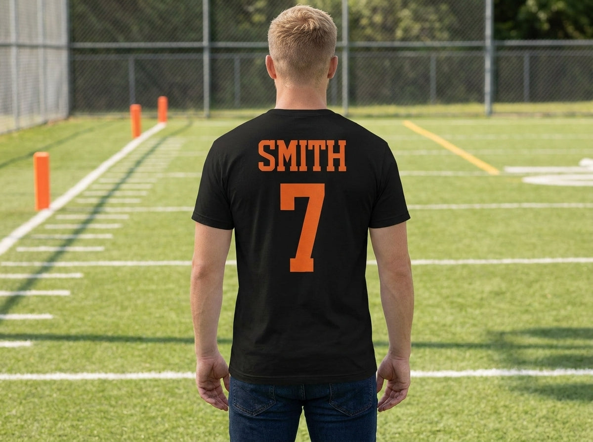 A man viewed from behind wearing a black t-shirt with 'SMITH' and the number '7' printed in orange on the back, standing on an outdoor sports field.