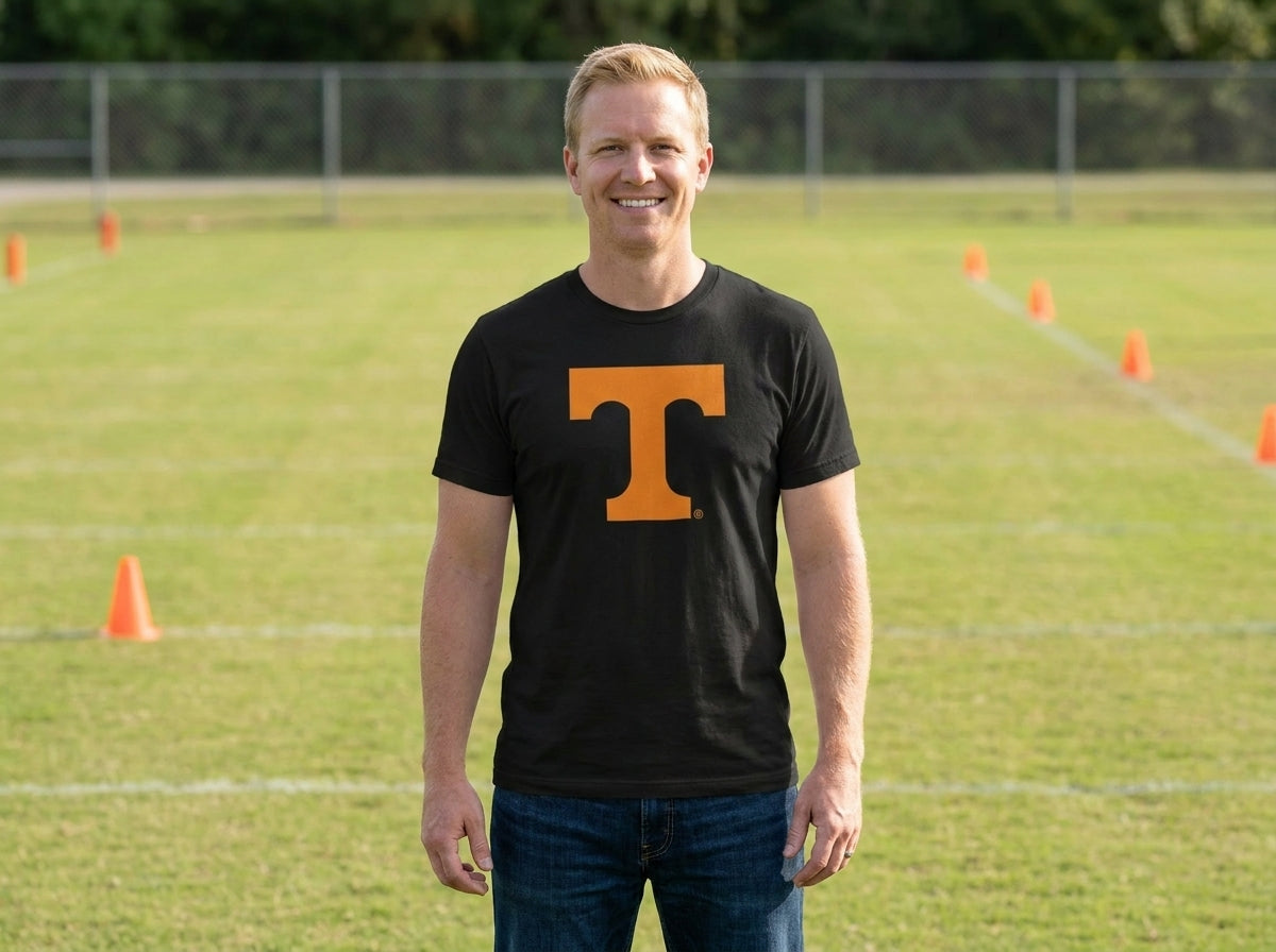 A smiling man standing on a grassy athletic field wearing a black short-sleeve t-shirt with a large orange block letter 'T' on the chest, paired with blue jeans.