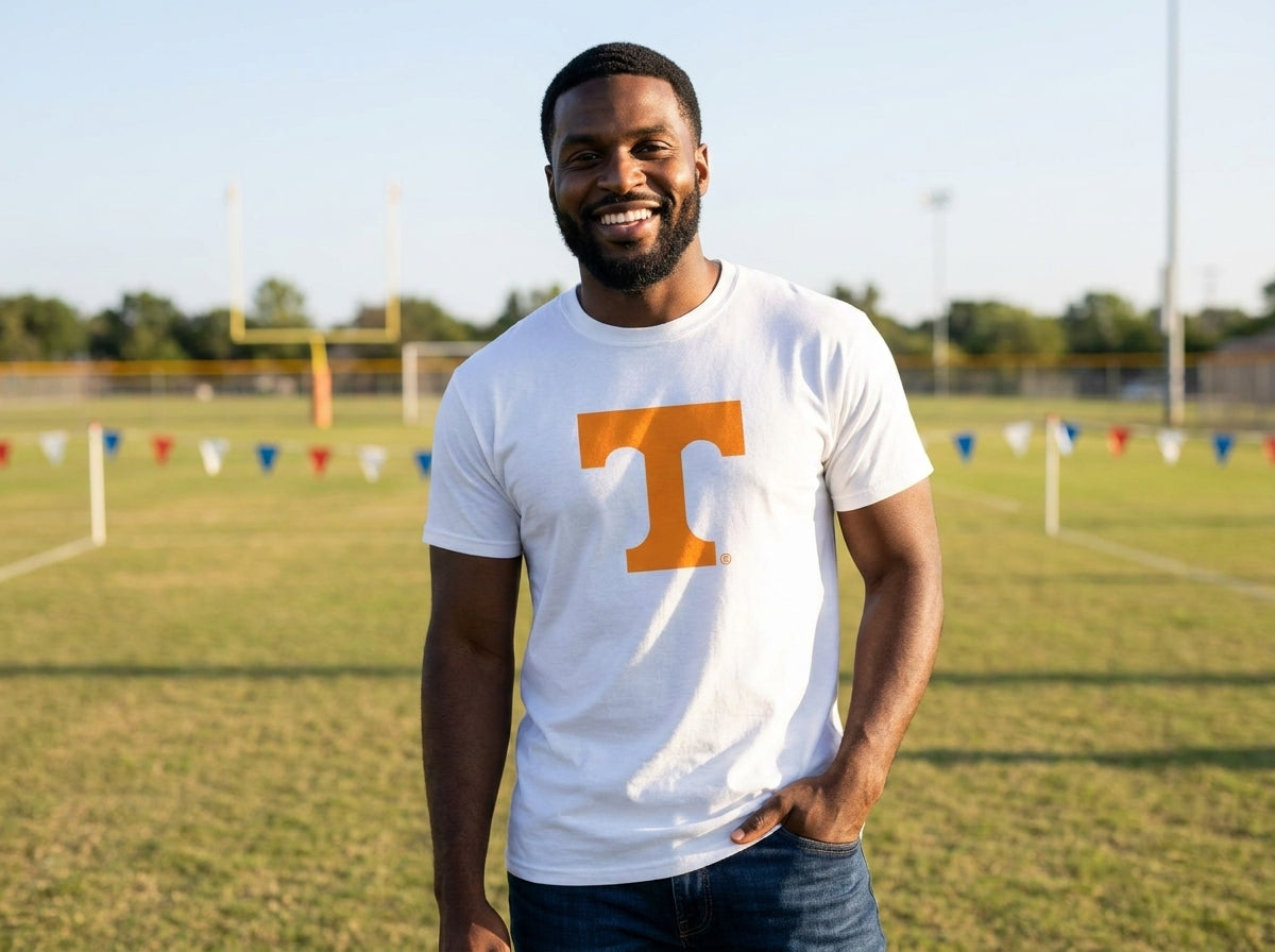 A smiling man standing on a grassy sports field wearing a white crewneck t-shirt with a large orange block letter 'T' on the chest, paired with dark blue jeans.