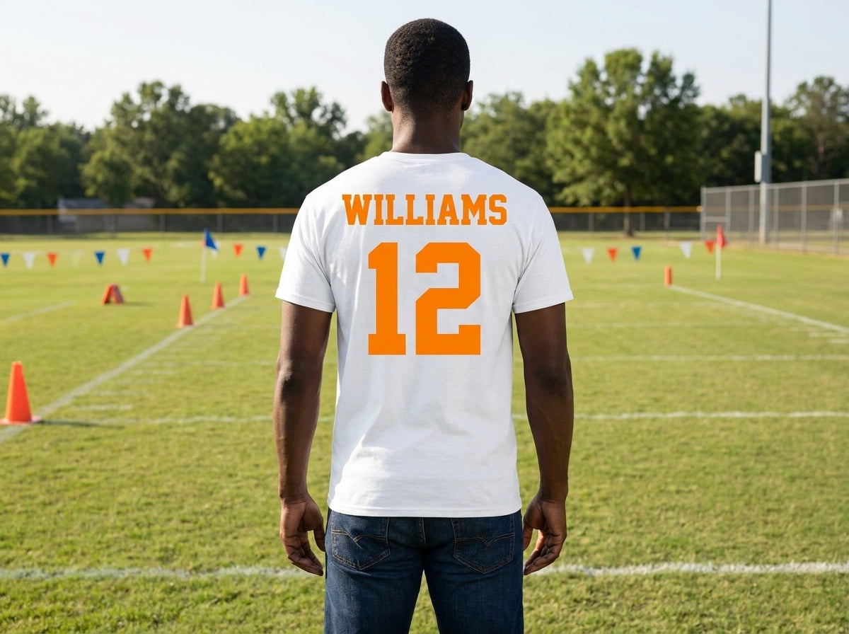 A man viewed from behind wearing a white t-shirt with 'WILLIAMS' and the number '12' printed in orange on the back, standing on a grassy football field.