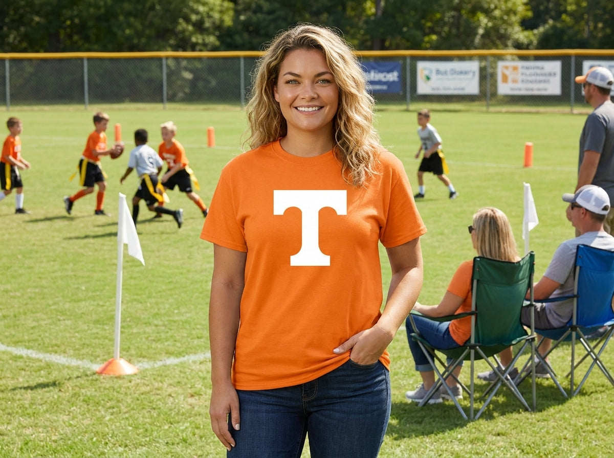 A woman standing on a grassy sports field wearing an orange t-shirt with a large white letter 'T' on the front, with children playing flag football in the background.