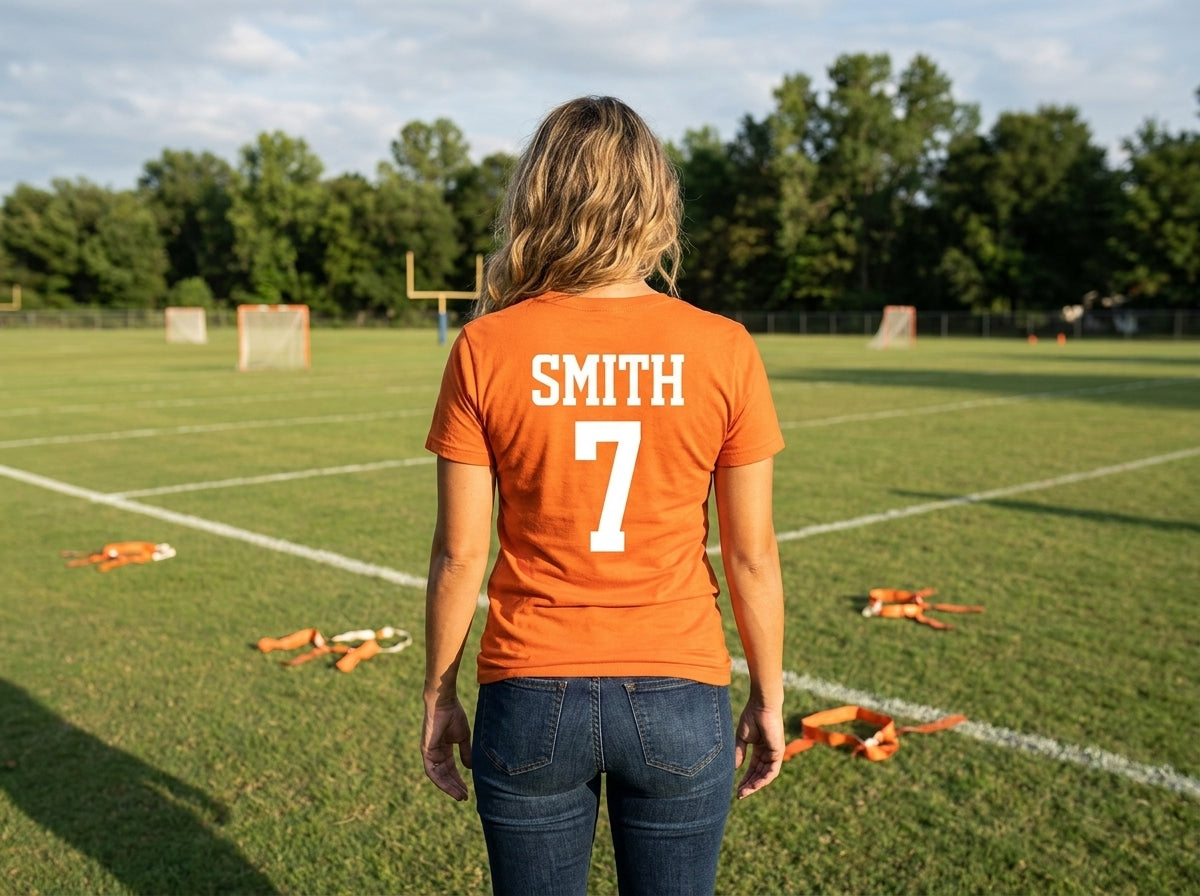 A woman viewed from behind stands on a grassy sports field wearing an orange short-sleeve T-shirt with the name 'SMITH' and number '7' printed in white on the back, paired with dark blue jeans.