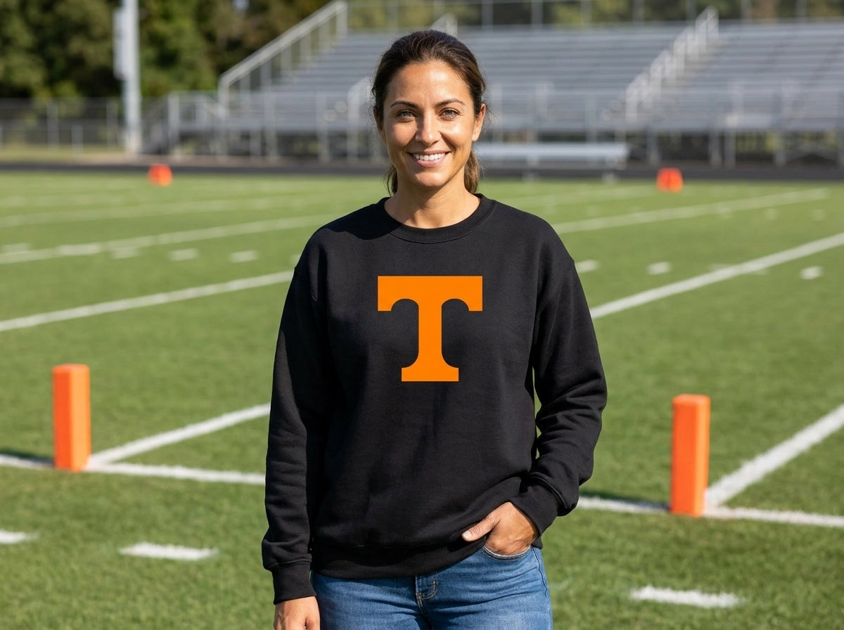 A woman stands on a football field wearing a black crewneck sweatshirt with a large orange letter 'T' on the chest, paired with blue jeans.