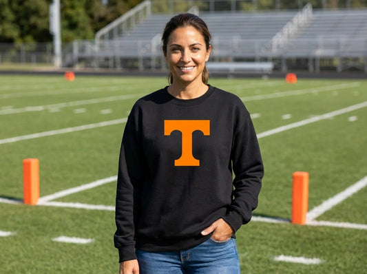 A woman stands on a football field wearing a black crewneck sweatshirt with a large orange letter 'T' on the chest, paired with blue jeans.