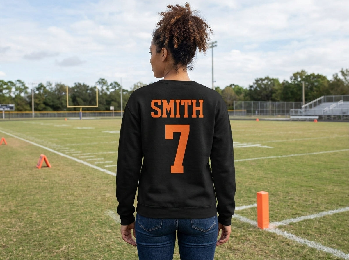 A woman stands on a football field wearing a black sweatshirt from the back, which features the name 'SMITH' and number '7' printed in orange. She is also wearing blue jeans, with the field, trees, and sky visible in the background.