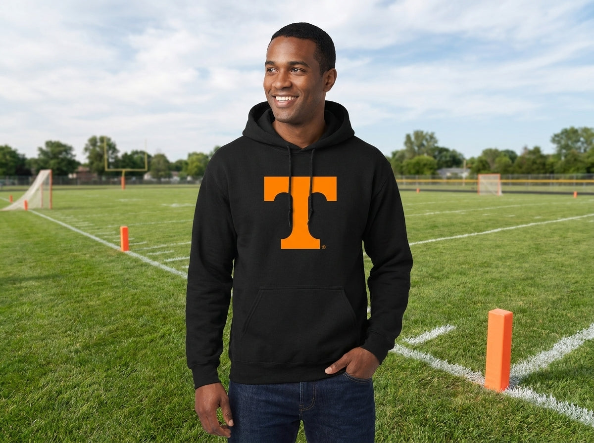 A man standing on a grassy football field wearing a black pullover hoodie with a large orange block letter 'T' on the chest, paired with dark blue jeans.