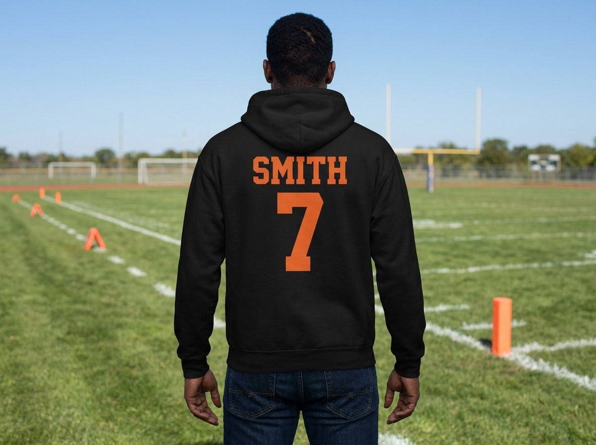 A man viewed from behind wearing a black hoodie with 'SMITH' and the number '7' printed in orange on the back, standing on a green football field under a blue sky.