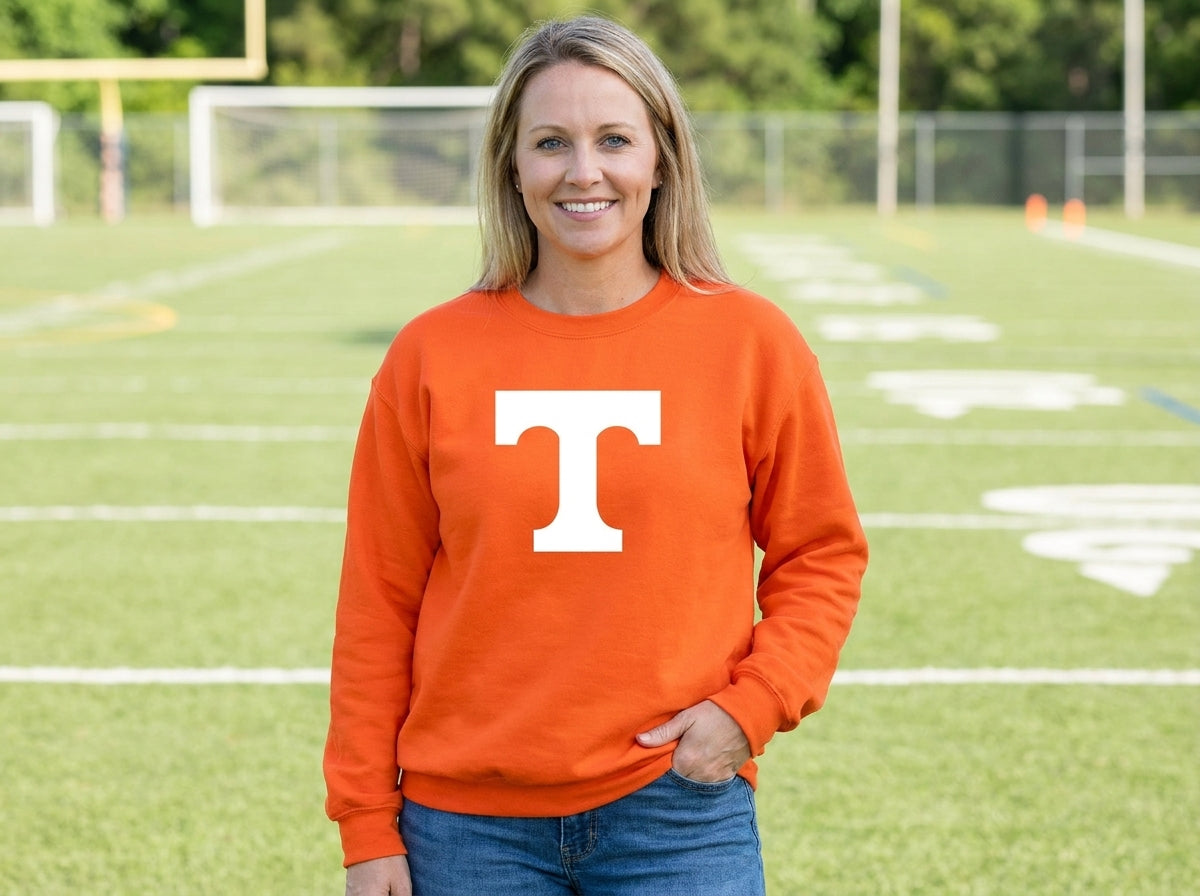 A woman stands on a football field wearing a orange crewneck sweatshirt with a large white 'T' logo on the front, paired with blue jeans.