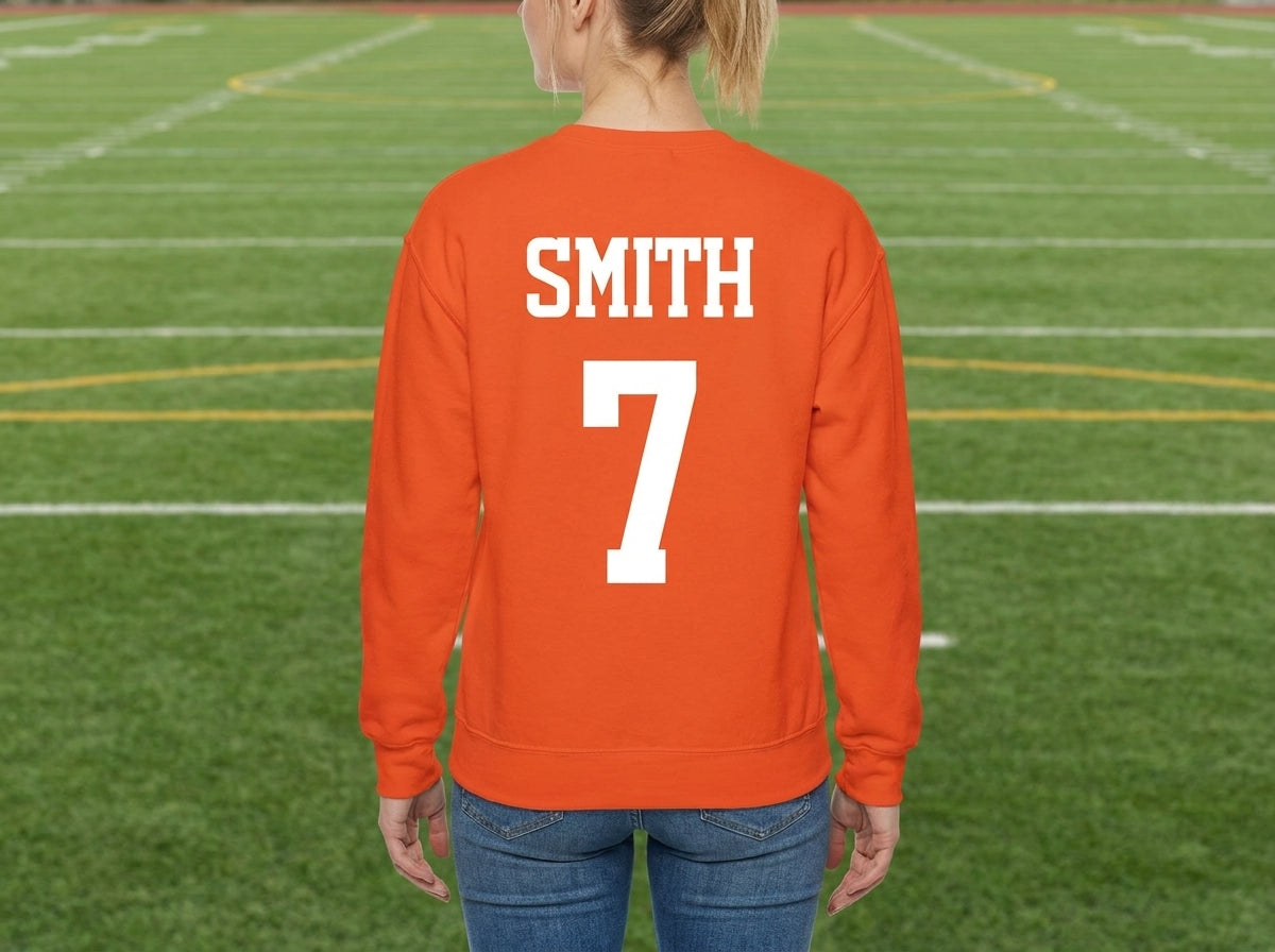 A woman viewed from behind wearing a orange sweatshirt with 'SMITH' and the number '7' printed in white on the back, standing on a green football field.