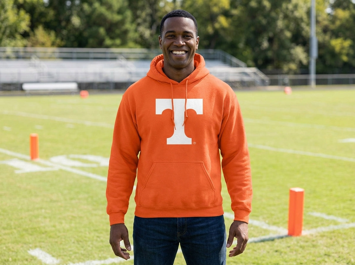 A smiling man standing on a football field wearing an orange hoodie with a large white 'T' logo on the front, paired with dark blue jeans.