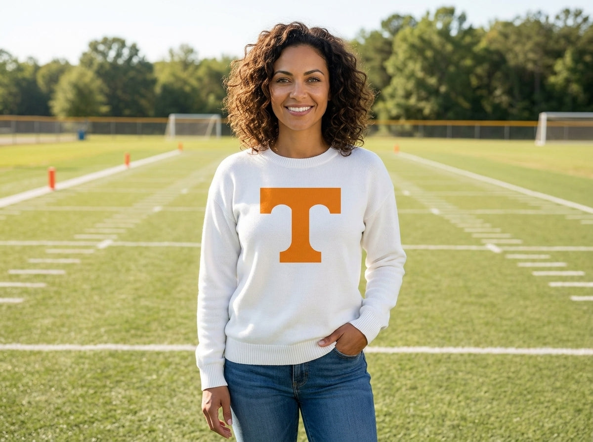 A woman stands on a football field wearing a white crewneck sweatshirt with a large orange 'T' logo on the front, paired with blue jeans.