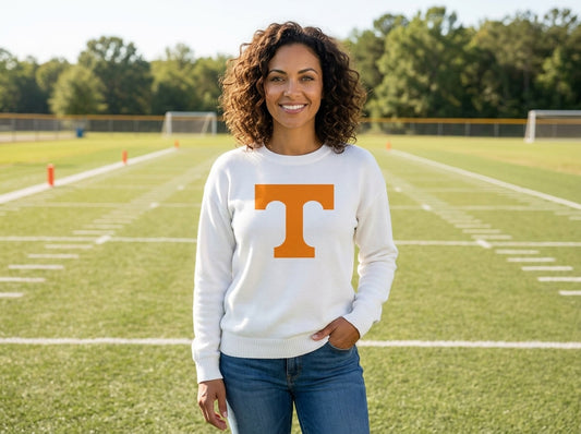 A woman stands on a football field wearing a white crewneck sweatshirt with a large orange 'T' logo on the front, paired with blue jeans.