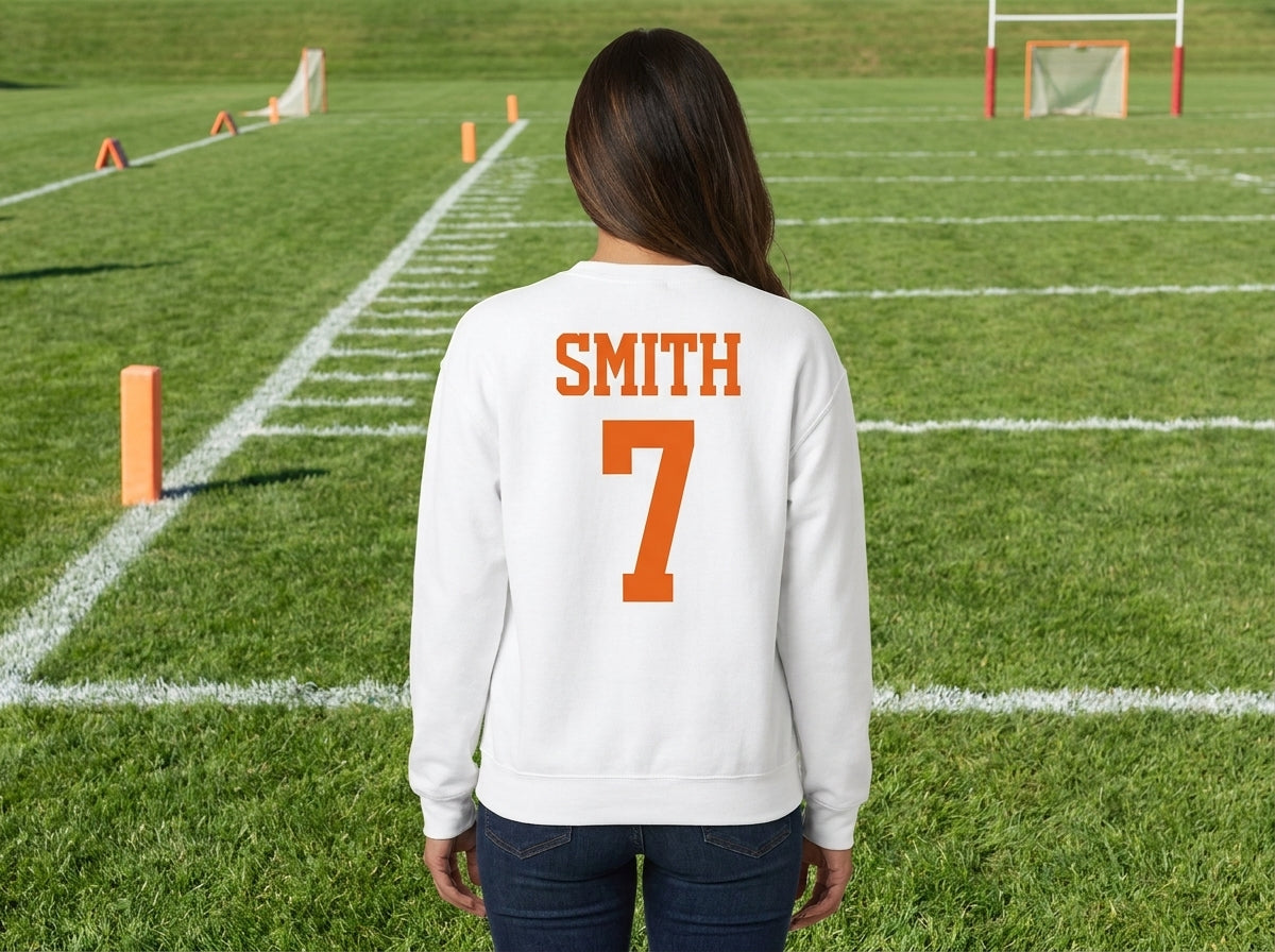 A woman viewed from behind wearing a white sweatshirt with 'SMITH' and the number '7' printed in orange on the back, standing on a green football field.