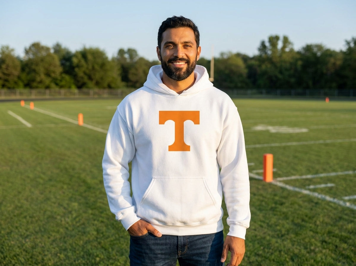 A smiling man wearing a white hoodie with a large orange letter 'T' on the chest, standing on a green football field at sunset.