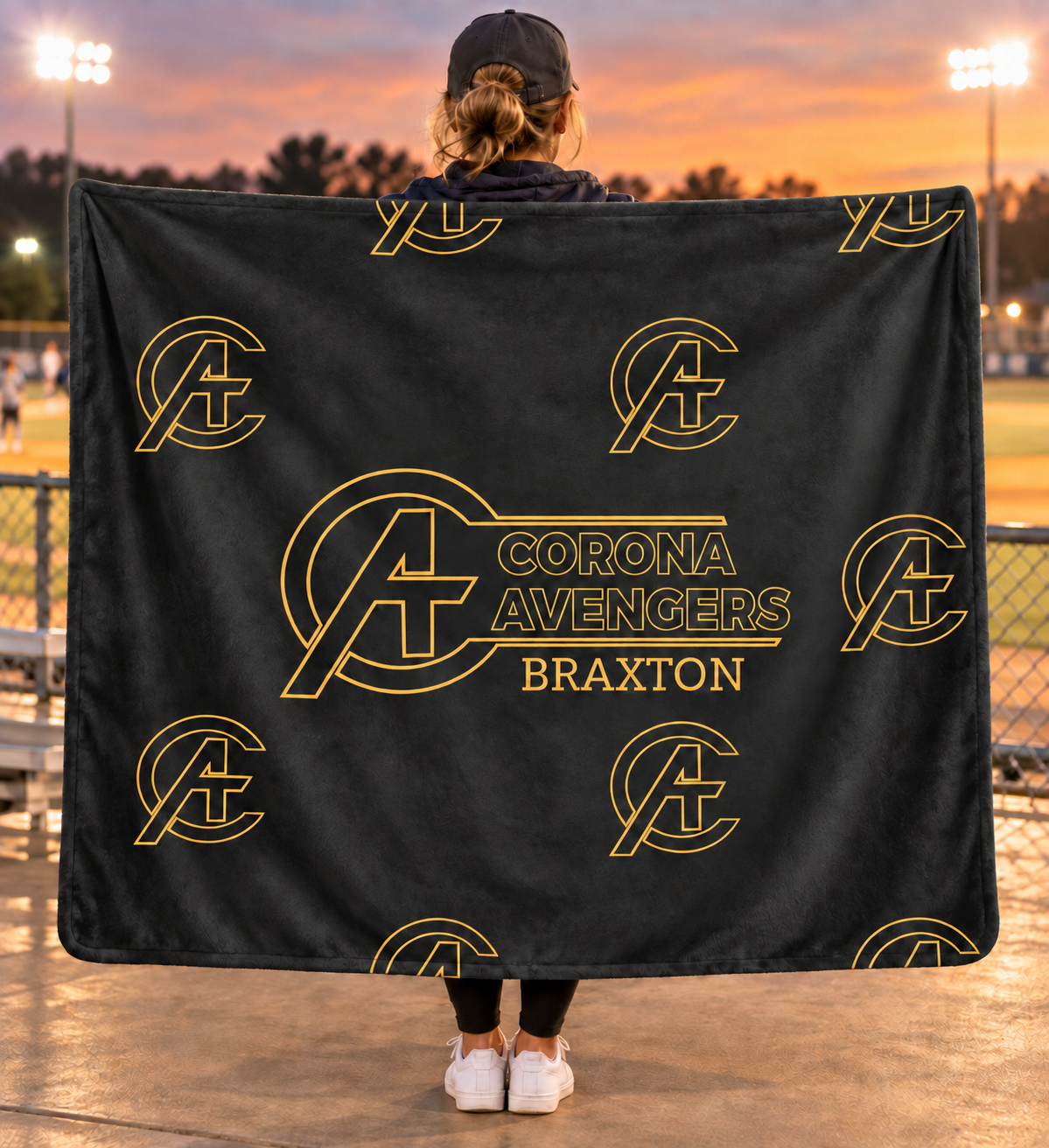 A person standing on a sports field at sunset holds up a large black blanket or throw featuring repeated gold Avengers-style logos and the text 'Corona Avengers Braxton' in gold. The person is shown from behind wearing a black cap, dark top, leggings, and white sneakers.