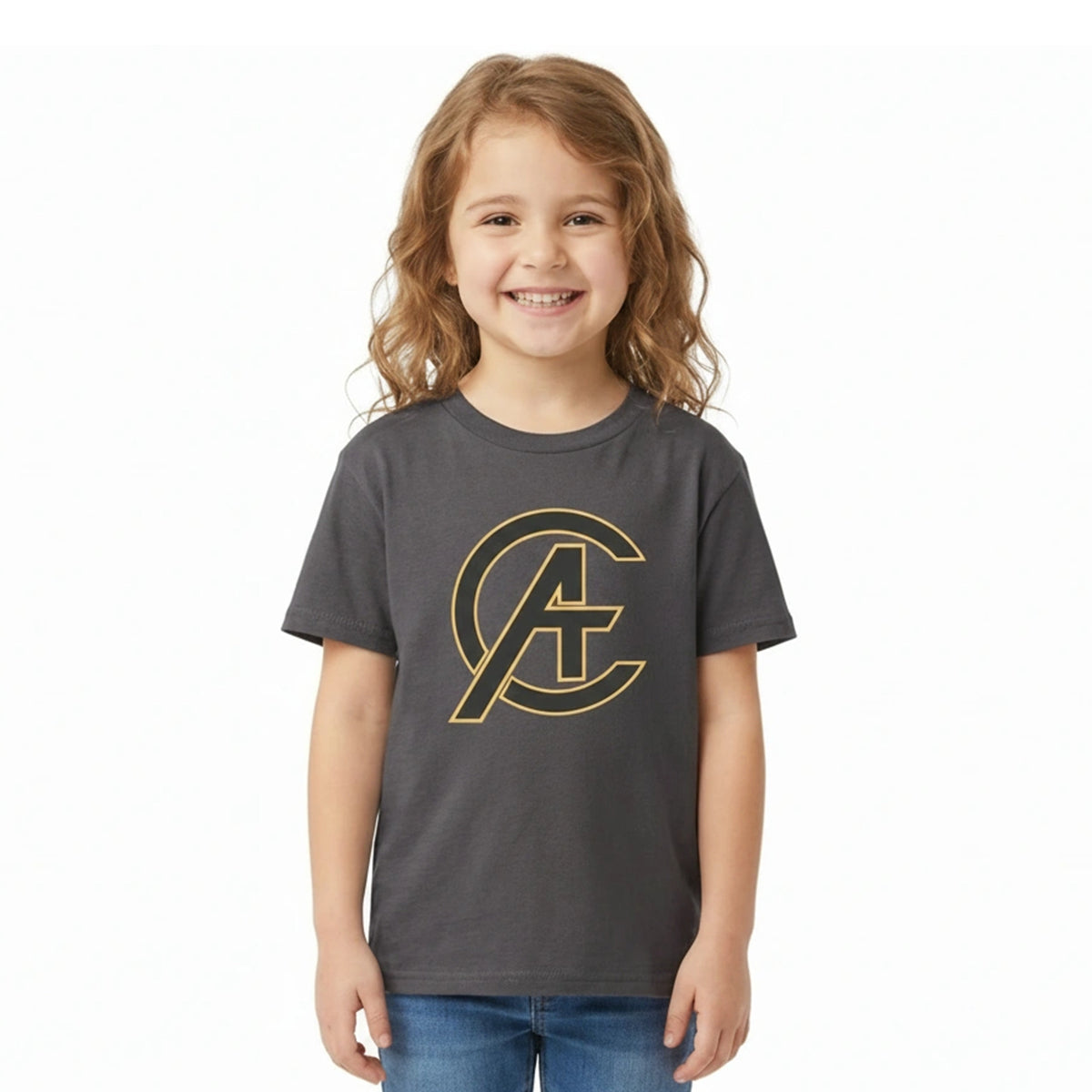 A young girl with wavy light brown hair smiling, wearing a dark gray t-shirt with a gold outline Avengers logo on the chest, paired with blue jeans, against a plain white background.