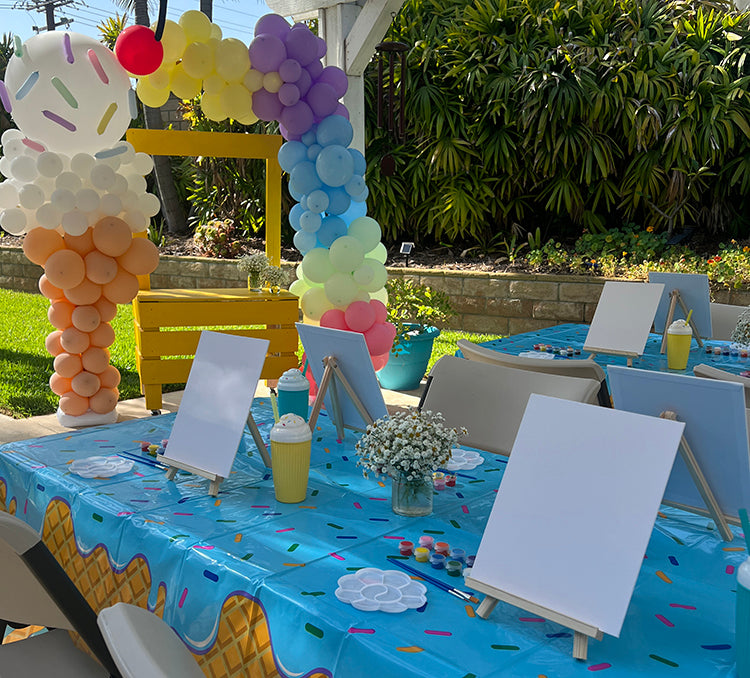 Decorated outdoor table with balloons, paint party art supplies, and a colorful tablecloth.