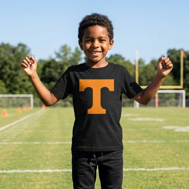 A young boy stands on a grassy football field wearing a black short-sleeve t-shirt with a large orange block letter 'T' on the chest, paired with black sweatpants. The background shows a sunny outdoor sports field with goalposts, trees, and a blue sky.