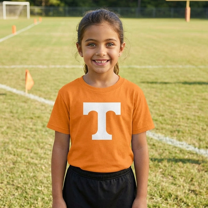 A young girl smiling on an outdoor sports field, wearing a bright orange short-sleeve t-shirt with a large white block letter 'T' on the chest, paired with a black athletic skirt.