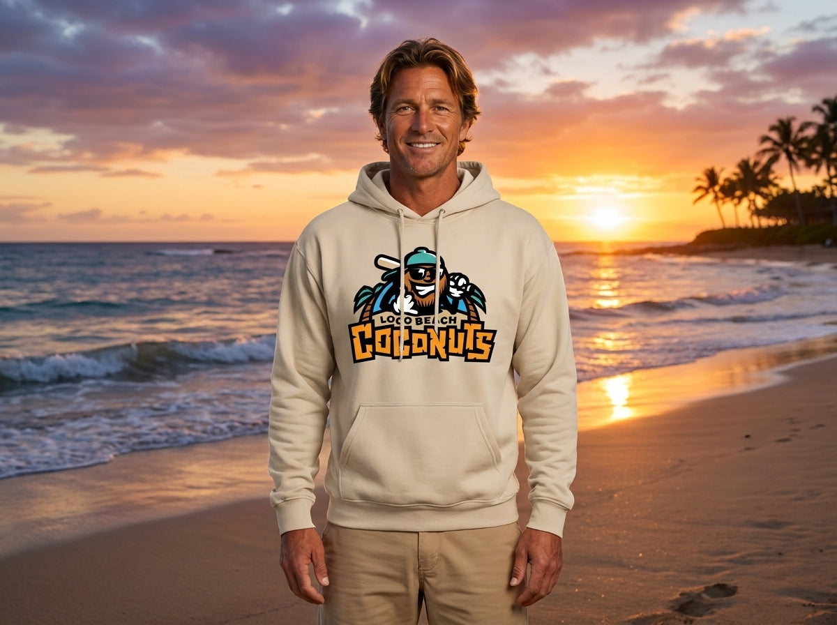 A man stands on a beach at sunset wearing a cream-colored hoodie with a colorful graphic on the chest, paired with khaki shorts.