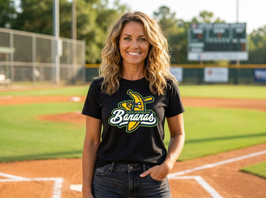 A smiling woman stands on a baseball field wearing a black graphic T-shirt paired with blue jeans.
