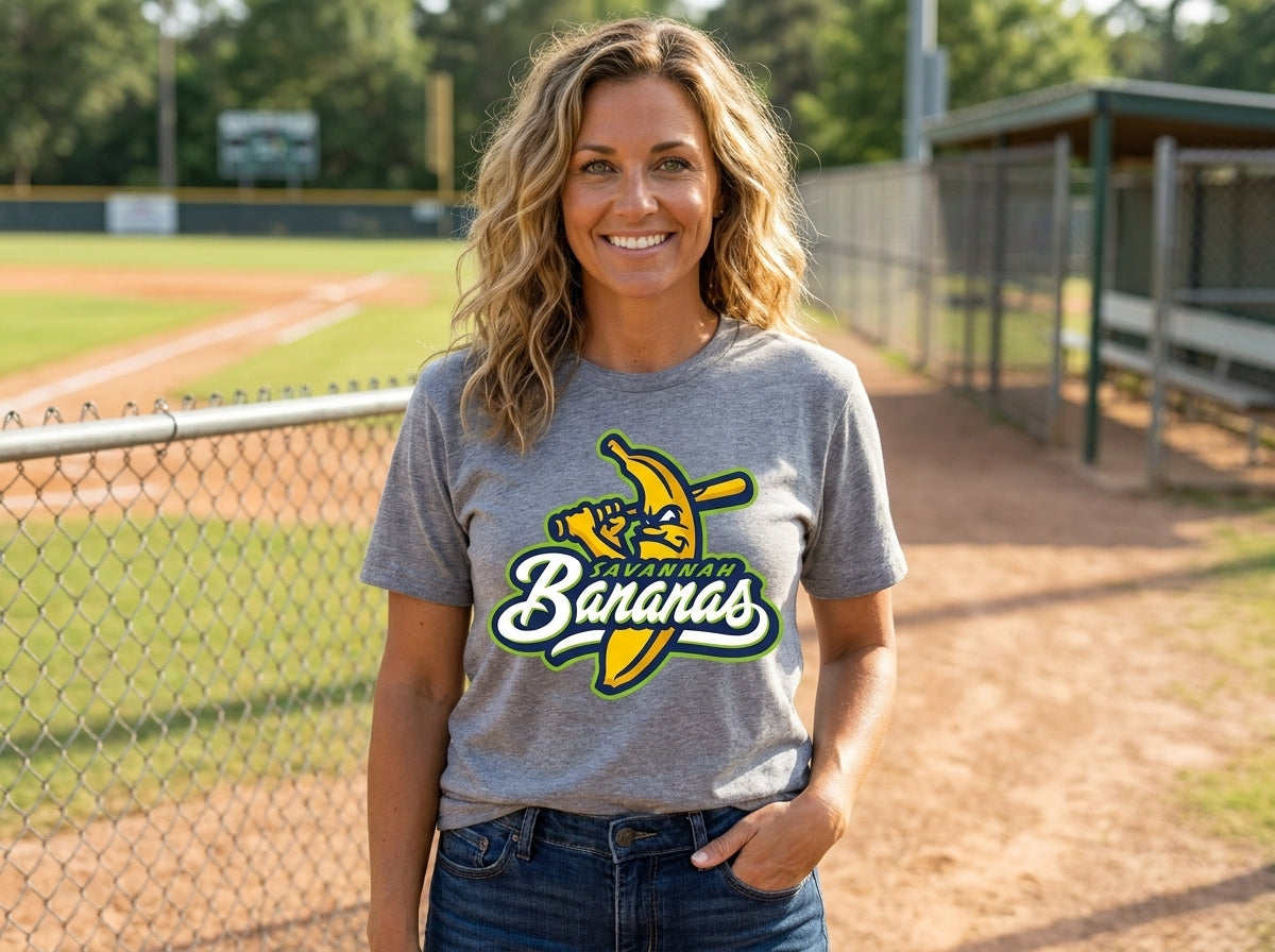 A smiling woman stands on a baseball field wearing a heather gray graphic T-shirt paired with blue jeans.
