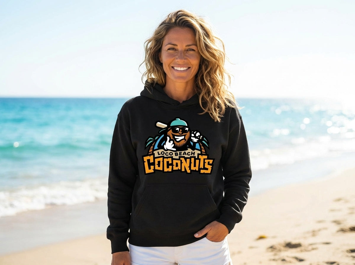 A woman stands on a sandy beach wearing a black hoodie with a colorful graphic on the front. The background shows the blue ocean and a bright sky.