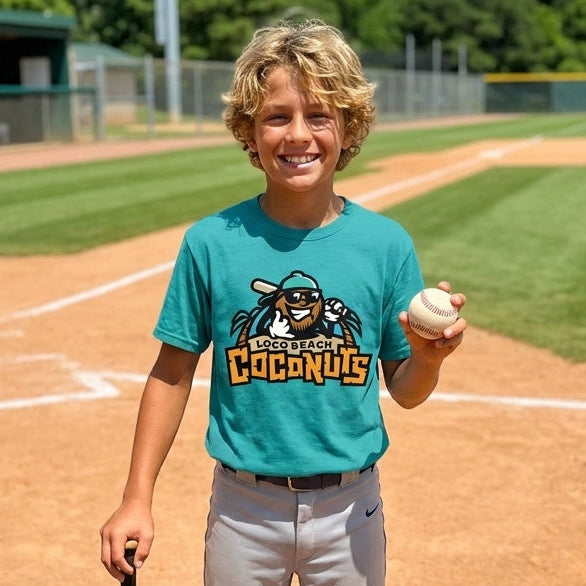 A young boy smiling on a baseball field wearing a teal graphic t-shirt that has custom logo, paired with gray pants and holding a bat and a baseball.