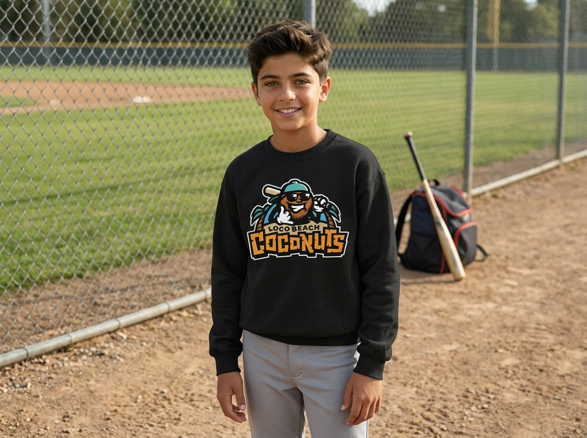A young boy stands on a baseball field wearing a black crewneck sweatshirt with a colorful cartoon logo and, paired with light gray pants. A chain-link fence, baseball field, and a bat bag are visible in the background.