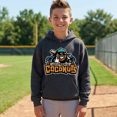 A young boy stands on a baseball field dirt path wearing a dark gray hoodie with a custom logo, gray sweatpants, and athletic shoes. A chain-link fence and baseball field are visible in the background.