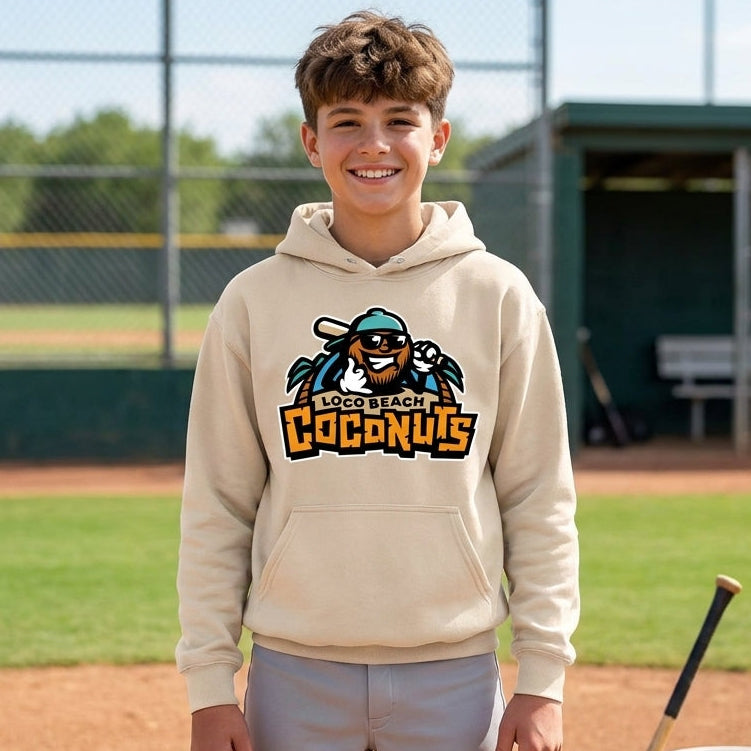 A smiling young man stands on a baseball field wearing a sand colored hoodie with a custom logo graphic, paired with gray sweatpants. A baseball bat and a bucket of balls are visible on the field next to him.