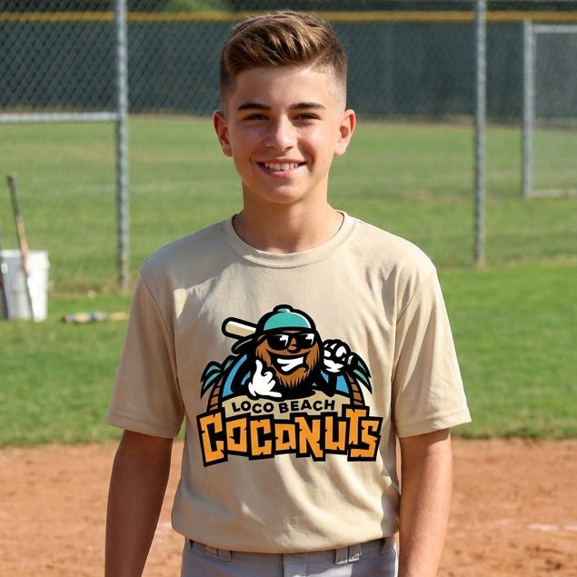 A young boy stands on a baseball field wearing a beige short-sleeve t-shirt with a logo graphic and gray baseball pants. The background shows a chain-link fence, green grass, and a dirt home plate area.